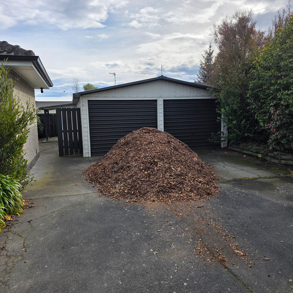A large pile of brown wood chips or mulch sits on a concrete driveway in front of a white garage with dark roll-up doors, with trees and another house visible in the background.