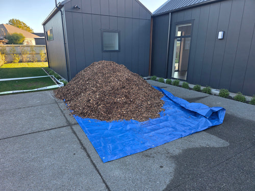 A large pile of brown wood chips or mulch sits on top of a bright blue tarp spread across a concrete driveway in front of a modern dark gray house.